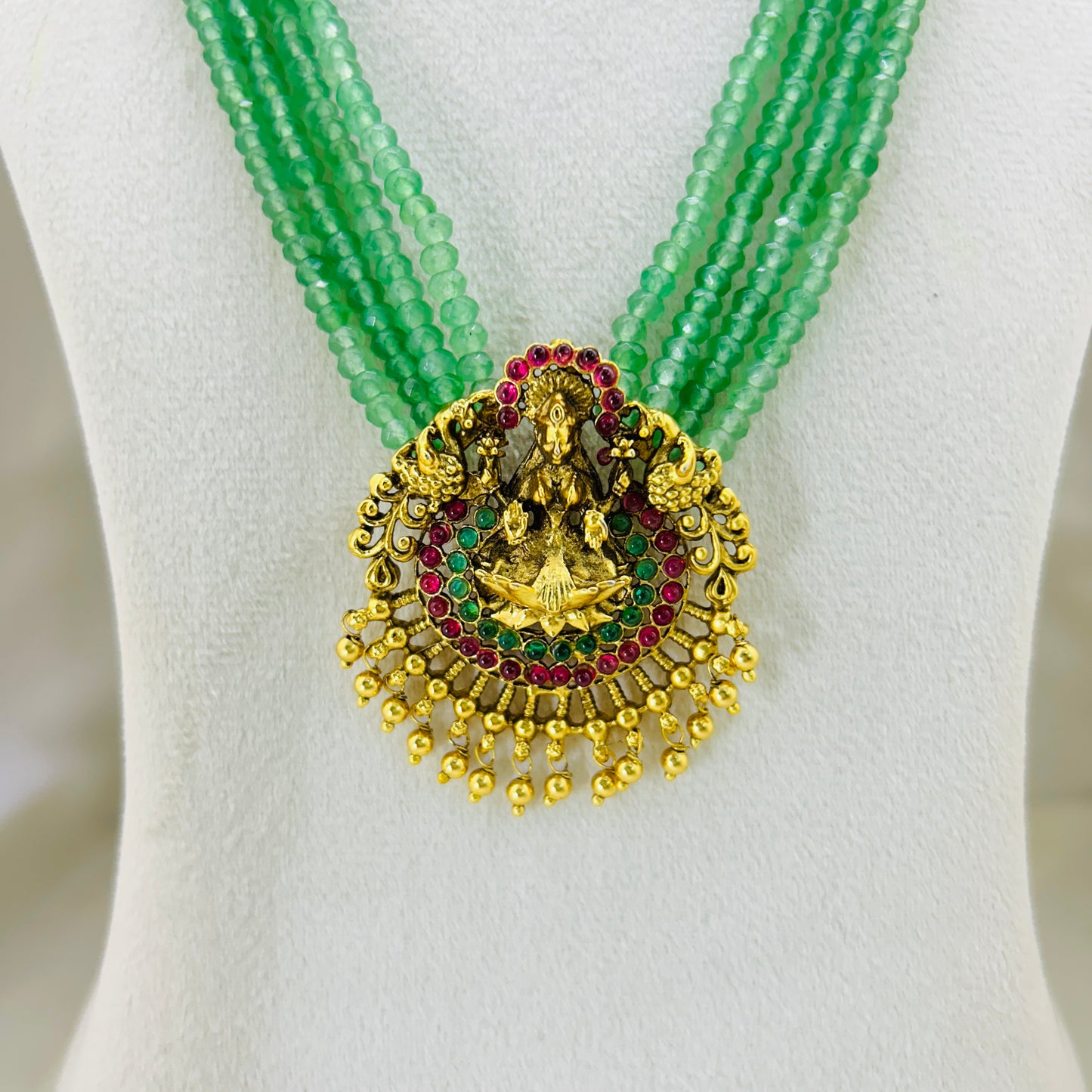 Green beaded necklace with a gold and gemstone pendant on a white mannequin.