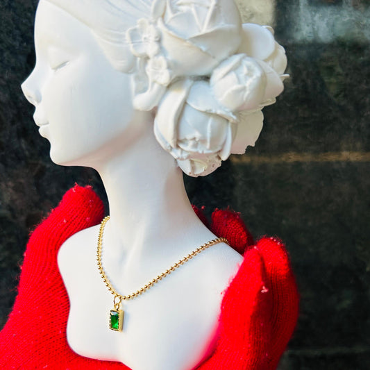 Mannequin head with white hair styled in a bun, wearing red gloves and a gold necklace with a green gemstone.